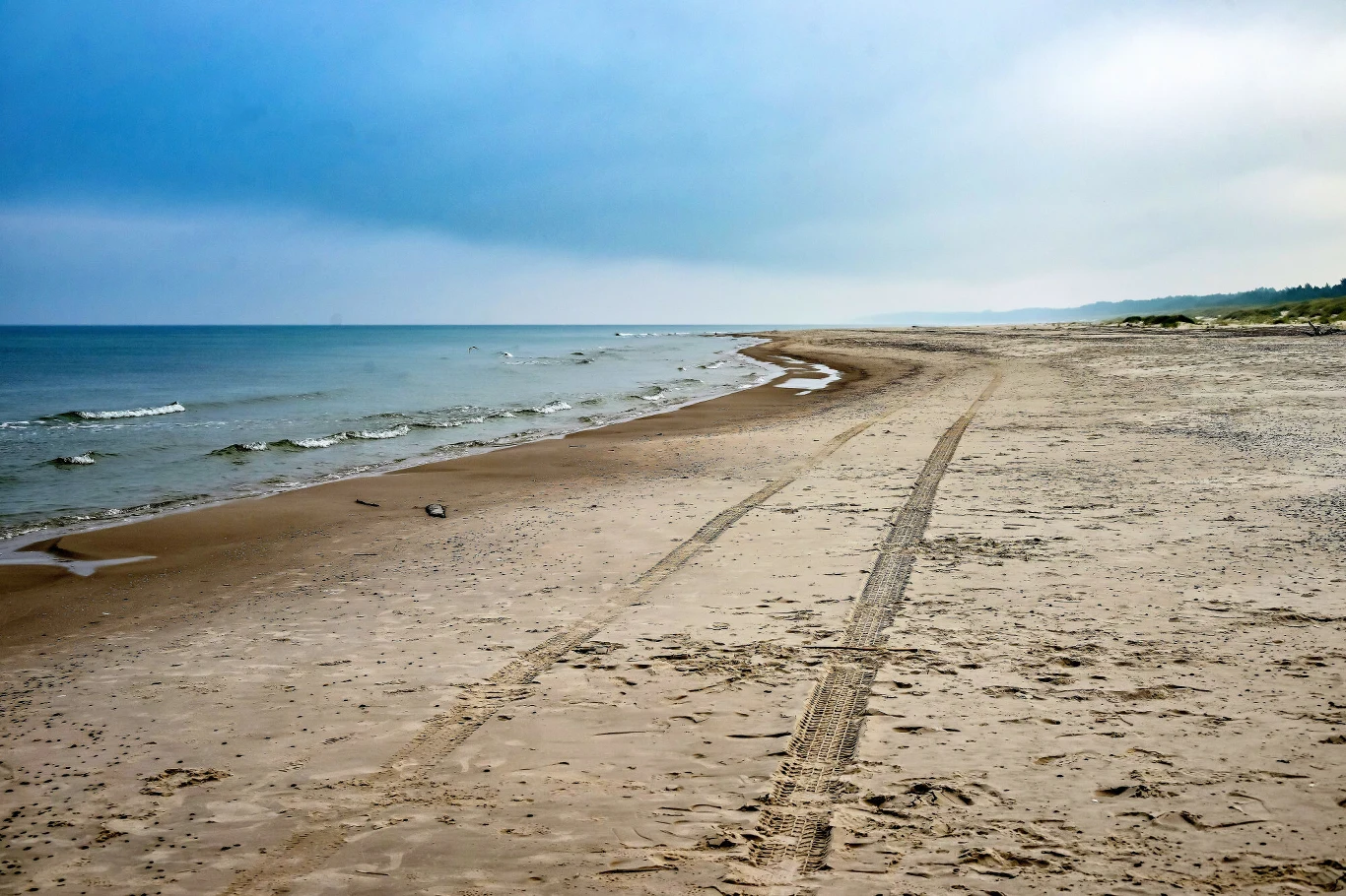 Szeroka, piaszczysta plaża z śladami opon prowadzącymi wzdłuż linii brzegu, spokojne morze i delikatnie zachmurzone niebo nad horyzontem.