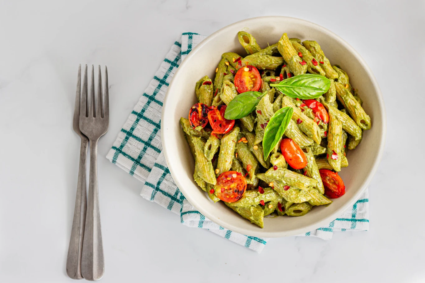 Makaron penne z pesto bazyliowym, kawałkami pomidorów koktajlowych i świeżymi listkami bazylii, podany w misce na biało-zielonej serwetce obok dwóch widelców.
