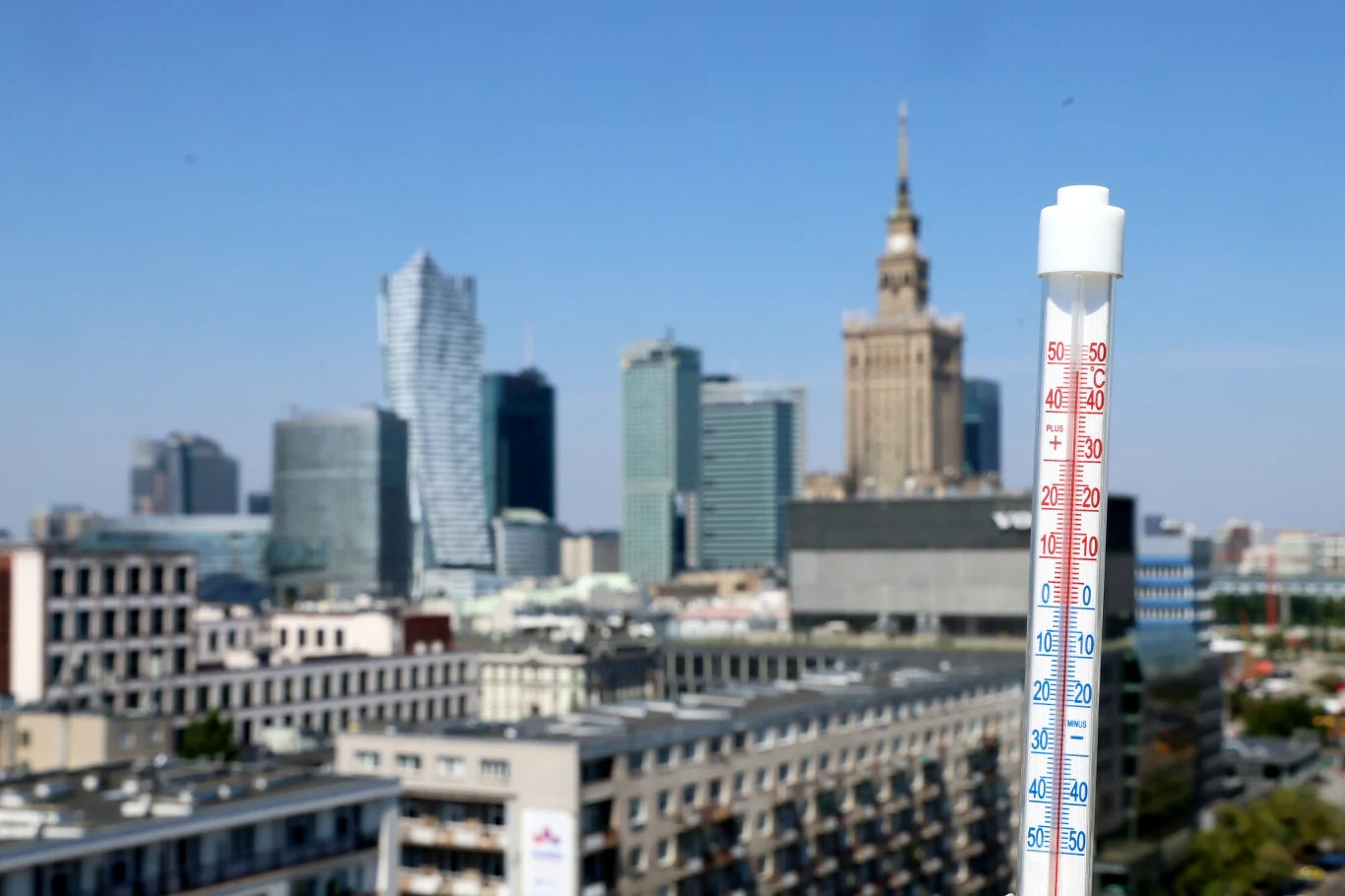 Termometr mierzący wysoką temperaturę na tle panoramy centrum Warszawy z widocznym Pałacem Kultury i Nauki oraz nowoczesnymi wieżowcami.