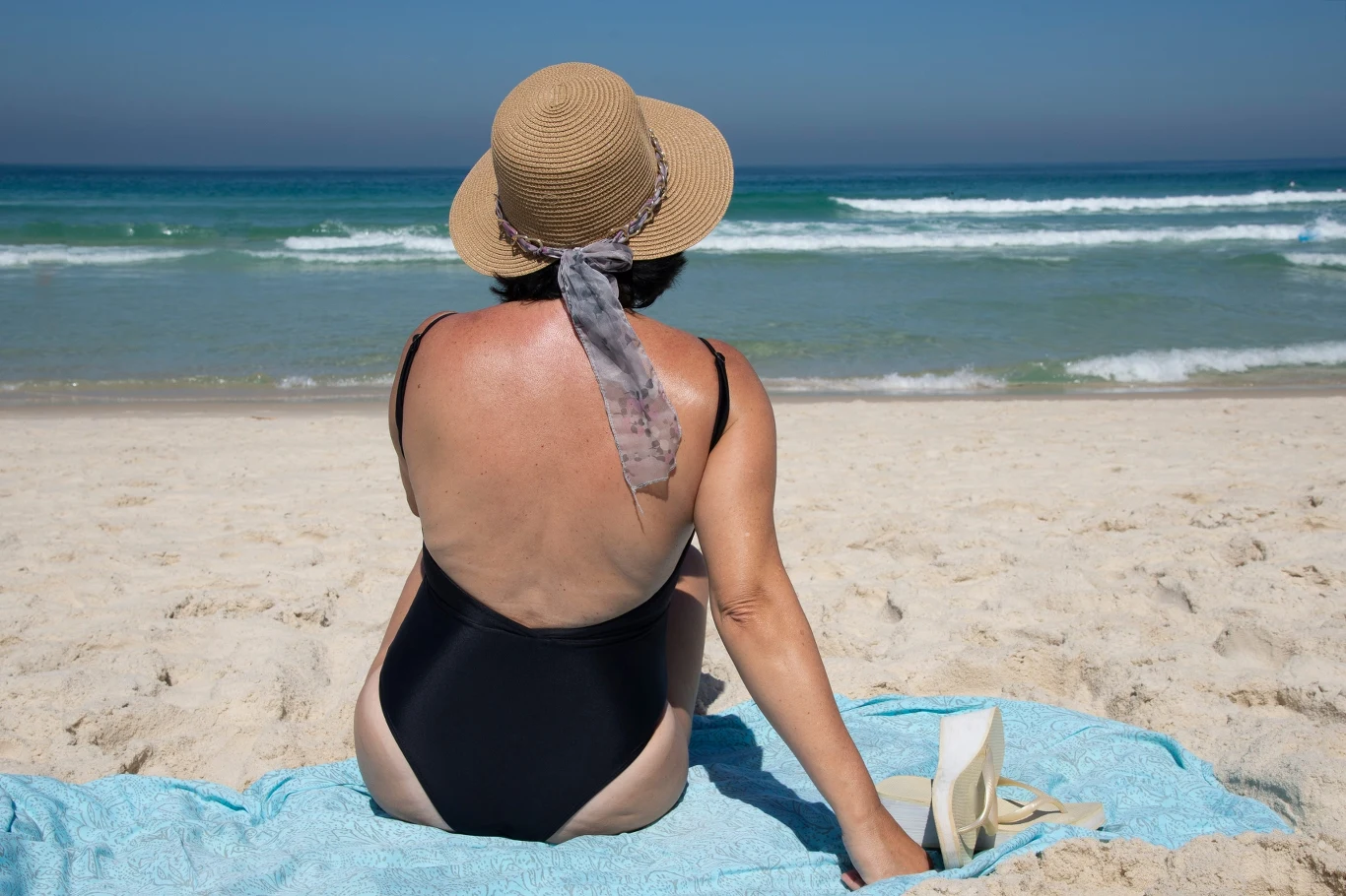 Kobieta w czarnym stroju kąpielowym i kapeluszu z szerokim rondem siedząca na niebieskim ręczniku na piaszczystej plaży, patrząca w stronę morza.