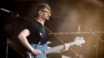 Mężczyzna w okularach przeciwsłonecznych gra na gitarze basowej i śpiewa do mikrofonu na scenie podczas koncertu.