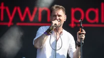 Mężczyzna w białej koszuli śpiewa do mikrofonu na scenie, w tle widoczne duże czerwone litery podświetlone na czarnym ekranie.