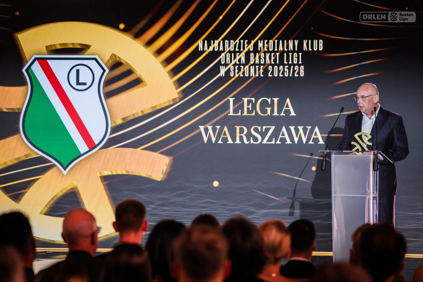 Mężczyzna w okularach przemawia na scenie przy mównicy, obok znajduje się duże logo Legii Warszawa oraz napis informujący, że klub został nagrodzony tytułem najbardziej medialnego klubu Orlen Basket Ligi w sezonie 2023/24.