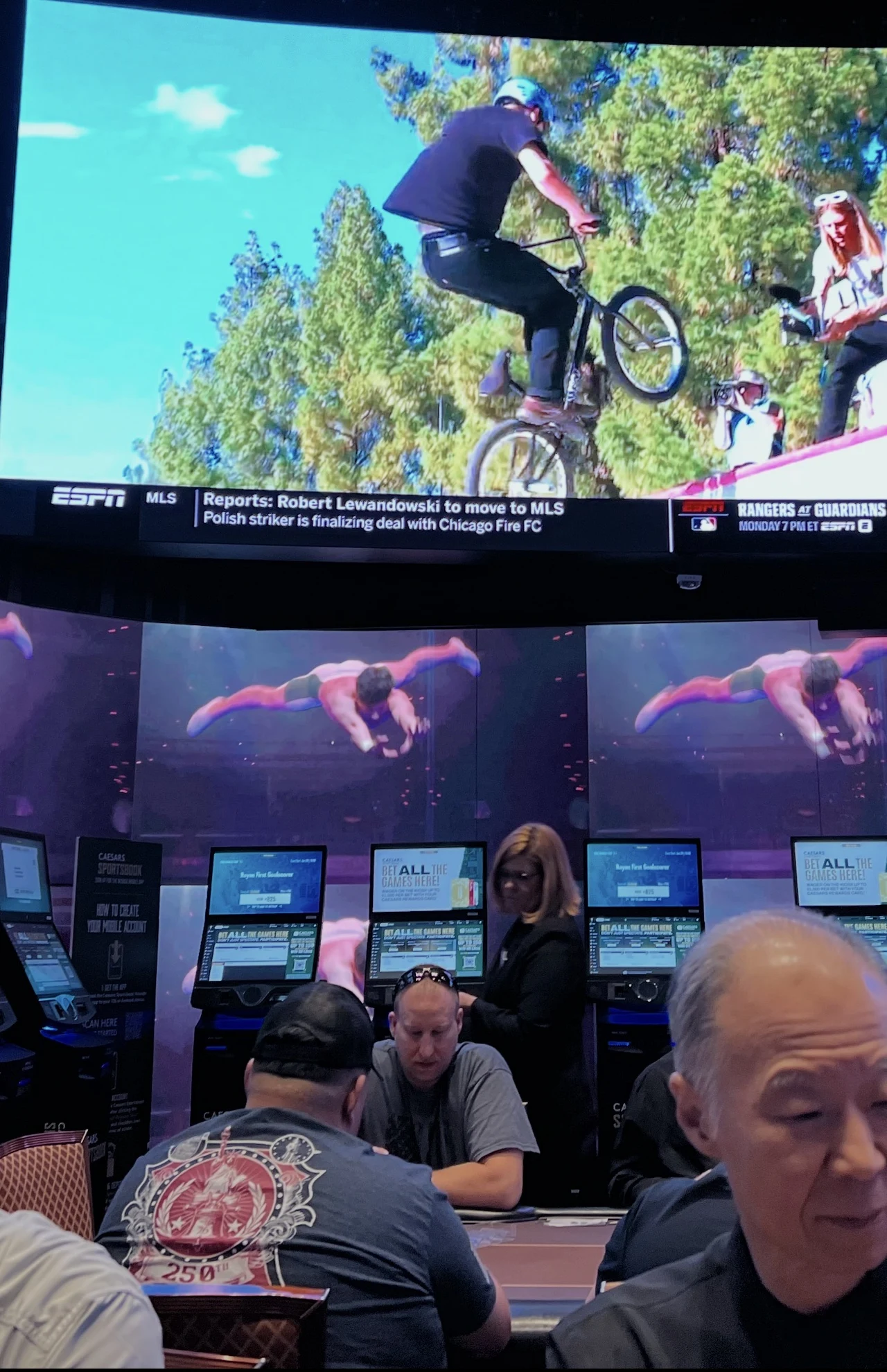 Stół do gry w kasynie z osobami skupionymi na rozgrywce, za nimi duży ekran wyświetlający dynamiczną scenę skoku rowerzysty BMX w powietrzu na tle drzew.