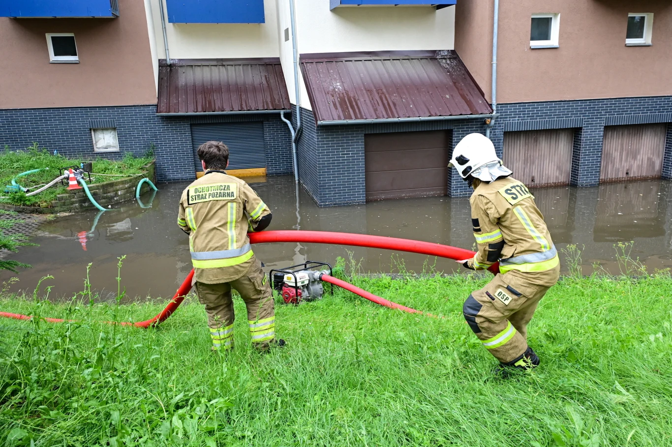Dwóch strażaków w mundurach przeciąga czerwony wąż strażacki na tle zalanych garaży w pobliżu budynków mieszkalnych.