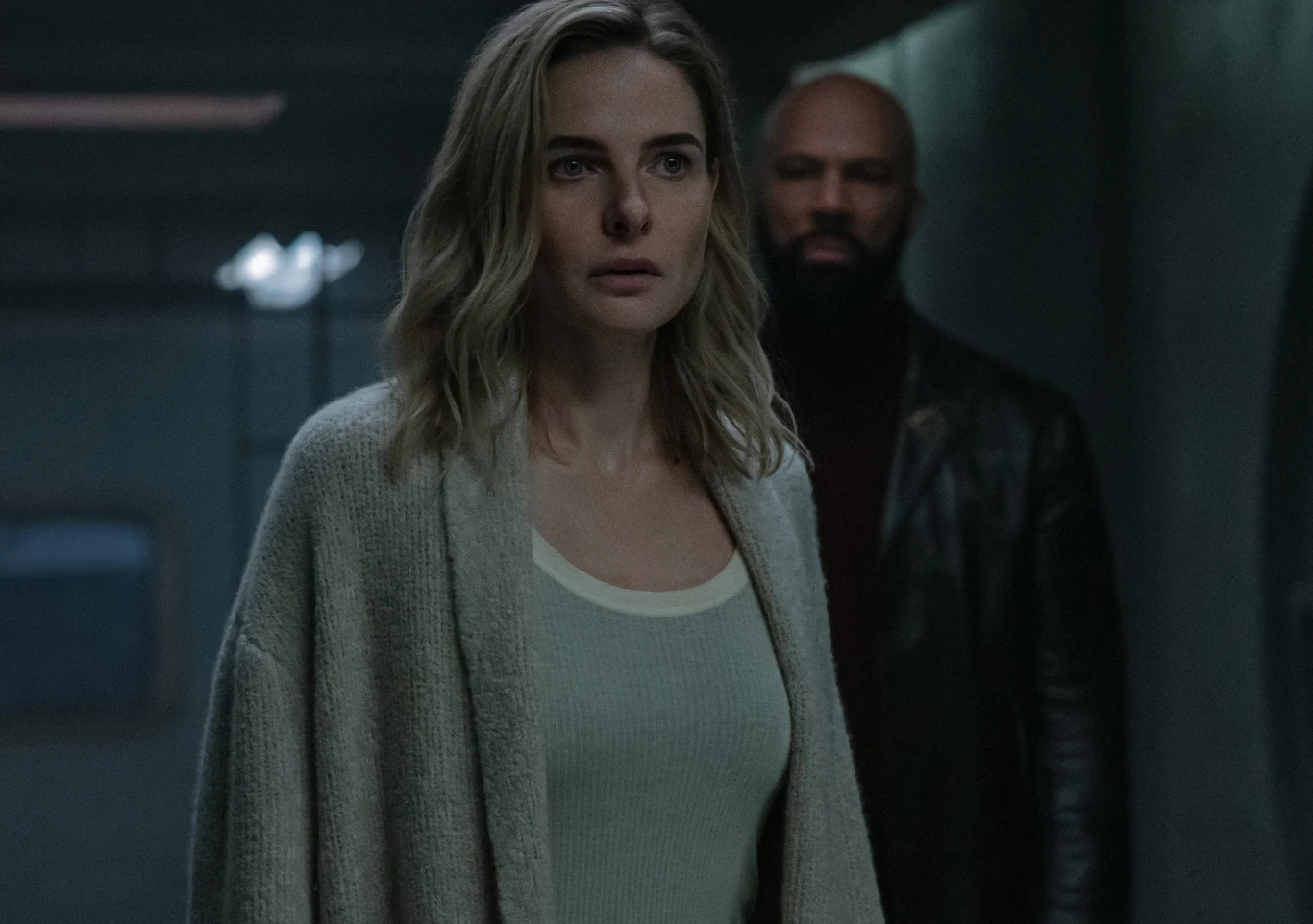 "Silos": Juliette (Rebecca Ferguson), Simms (Common) Kobieta o jasnych włosach ubrana w szary sweter stoi na pierwszym planie w słabo oświetlonym pomieszczeniu, za nią znajduje się mężczyzna z brodą ubrany w czarną kurtkę.