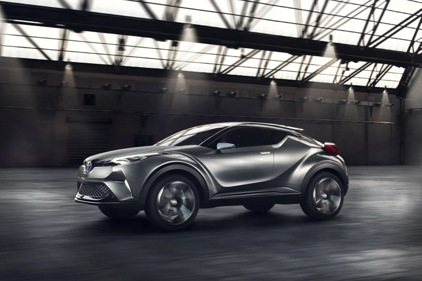 Futurystyczny srebrny SUV o dynamicznych liniach nadwozia na tle przestronnej, industrialnej hali z przeszkleniami w dachu.