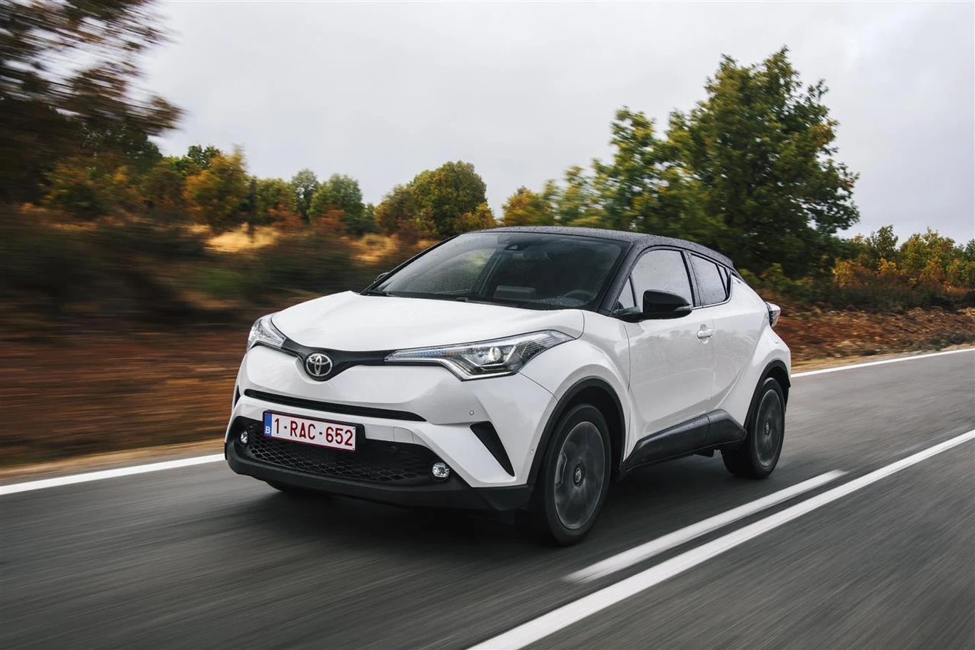 Toyota C-HR zadebiutowała w 2016 r. Biały samochód Toyota C-HR jedzie po dwupasmowej drodze otoczonej zielenią i jesiennymi drzewami.