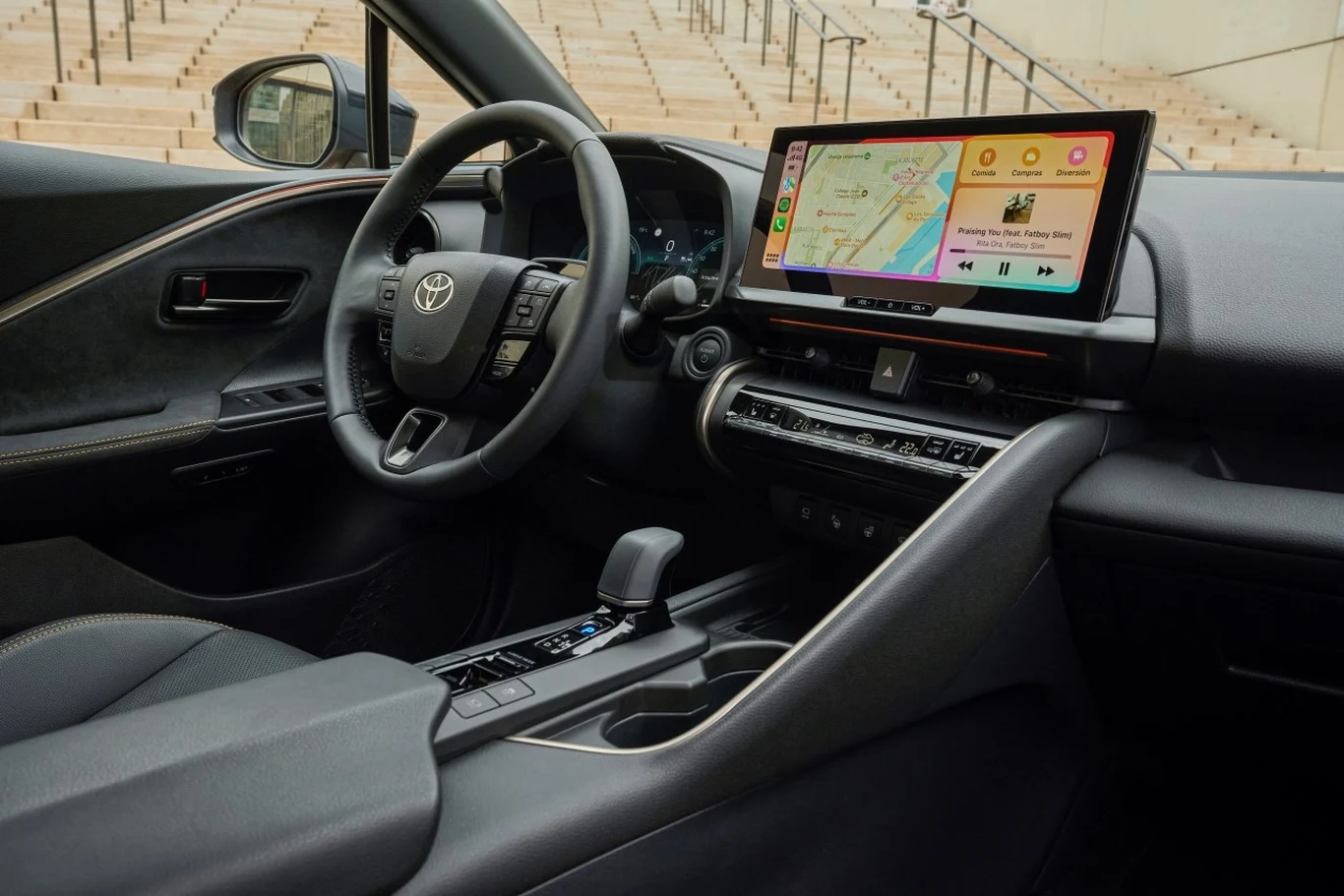 Wnętrze samochodu marki Toyota z cyfrowym wyświetlaczem na desce rozdzielczej, dużym ekranem multimedialnym z kolorową mapą i interfejsem, skórzaną kierownicą oraz nowoczesną konsolą centralną z automatyczną skrzynią biegów.