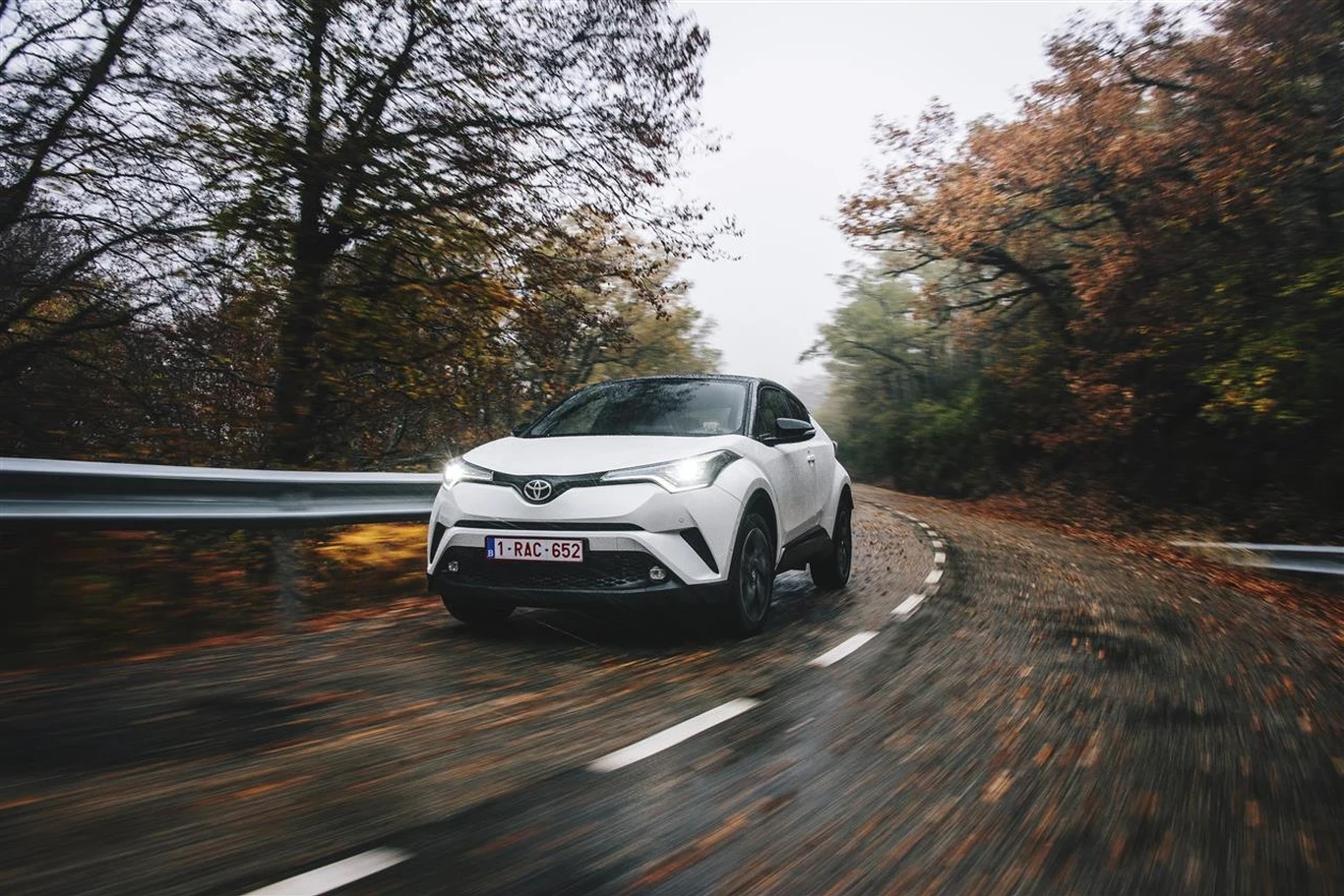 Białe auto marki Toyota jedzie krętą, wilgotną drogą otoczoną jesiennymi drzewami.