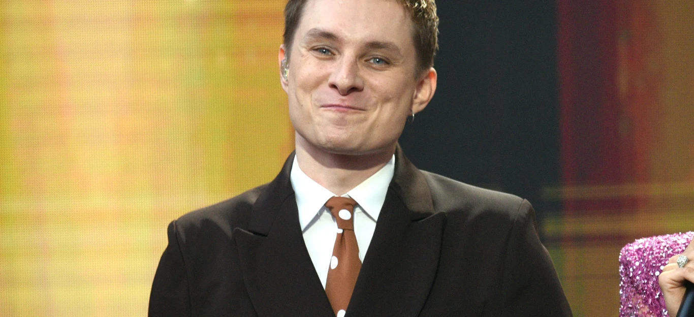 Ralph Kaminski Ralph Kaminski w ciemnej marynarce, białej koszuli i brązowym krawacie w białe kropki na tle jasnego światła.