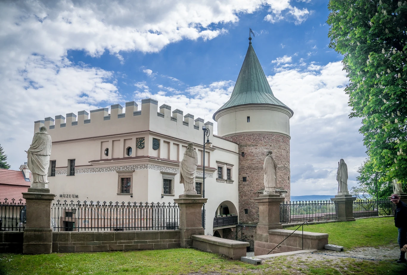 Zamek z cylindryczną wieżą i białą elewacją, otoczony kamiennym ogrodzeniem z rzeźbami postaci ustawionymi na cokołach, wokół zieleń i fragment brukowanej ścieżki.