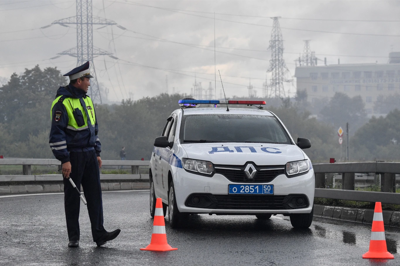 Policjant w mundurze stoi obok oznakowanego radiowozu z włączonymi sygnałami świetlnymi, a na jezdni ustawione są dwa pomarańczowe pachołki blokujące przejazd.