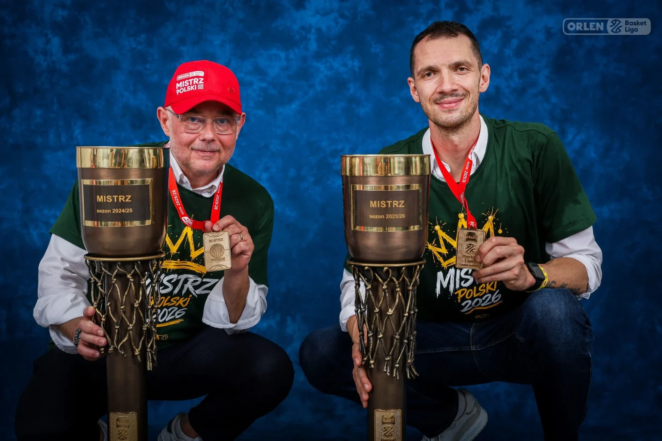 Dwóch mężczyzn w koszulkach mistrzowskich i z medalami na szyi trzyma duże trofea z napisem 'Mistrz Polski 2024' na tle niebieskiej ścianki z logo Orlen Basket Ligi.