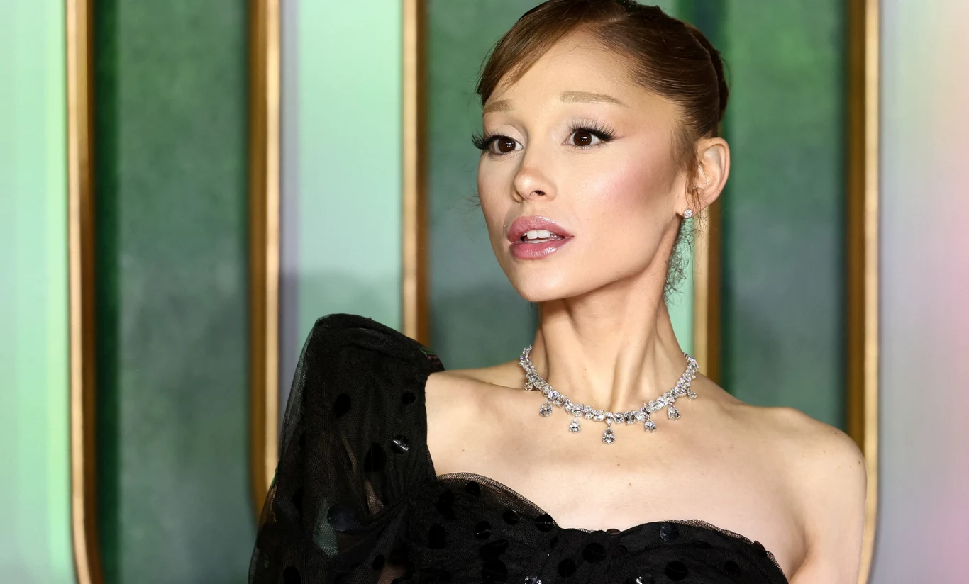 Ariana Grande Ariana Grande w czarnej sukni z odkrytym ramieniem, diamentową biżuterią, na tle zielono-złotych paneli.
