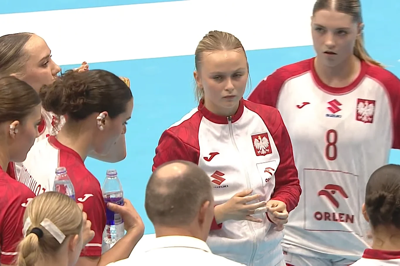 Zawodniczki reprezentacji Polski w piłce ręcznej ubrane w czerwono-białe stroje z orzełkiem na piersi zebrane wokół trenera podczas przerwy w meczu.