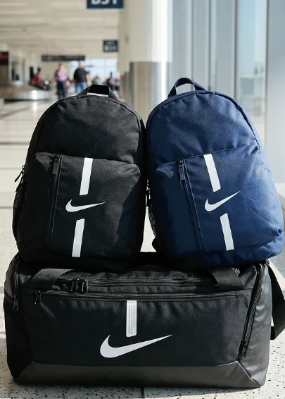 Dwa plecaki Nike, jeden czarny i jeden granatowy, ustawione na dużej czarnej torbie sportowej tej samej marki, w jasnym wnętrzu terminala lotniczego.