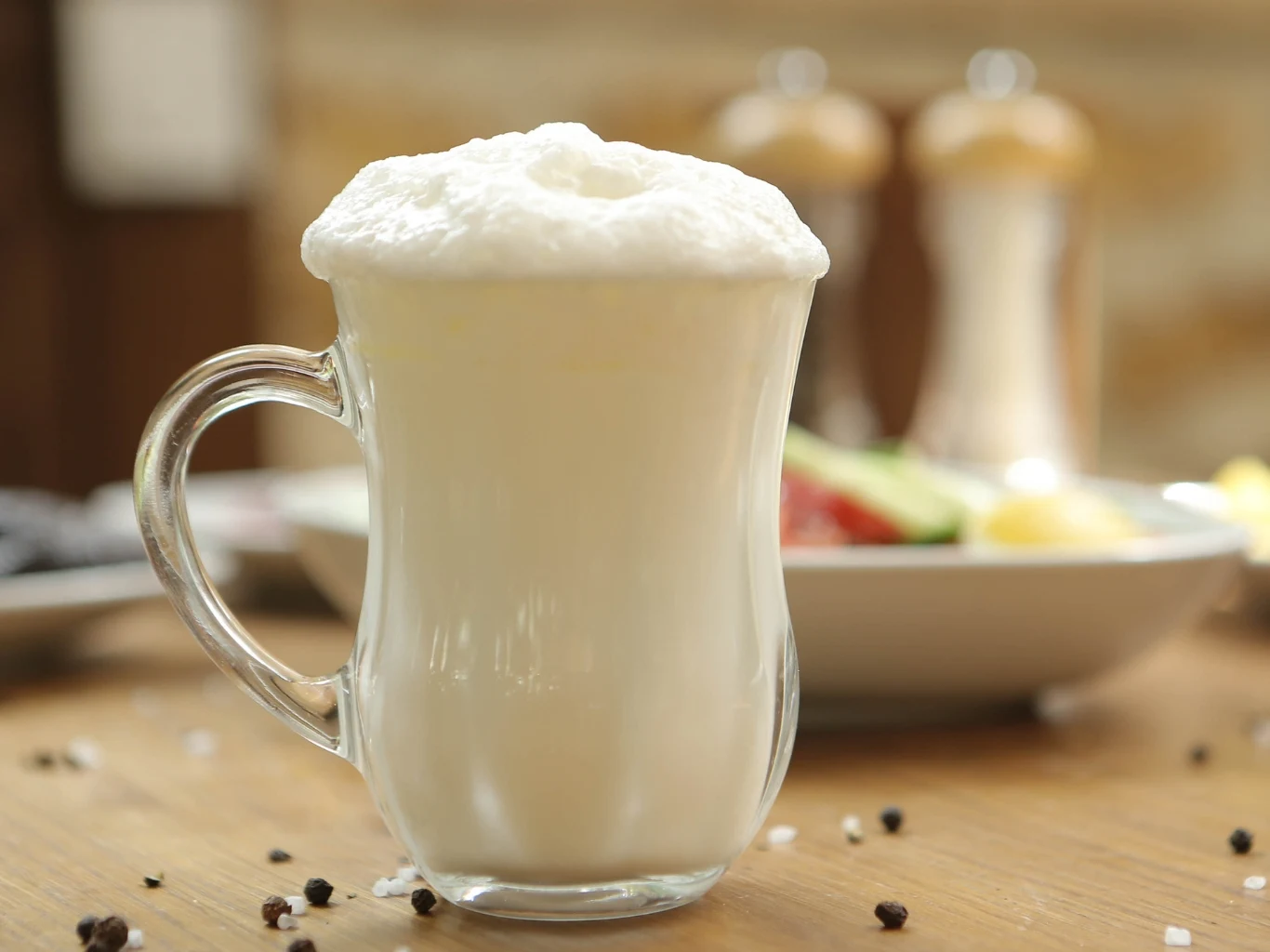 Ayran to turecki hit na upały. Nawadnia i gasi pragnienie Szklanka z ayranem o mlecznobiałej barwie i pianką, ustawiona na drewnianym stole obok talerza z jedzeniem.