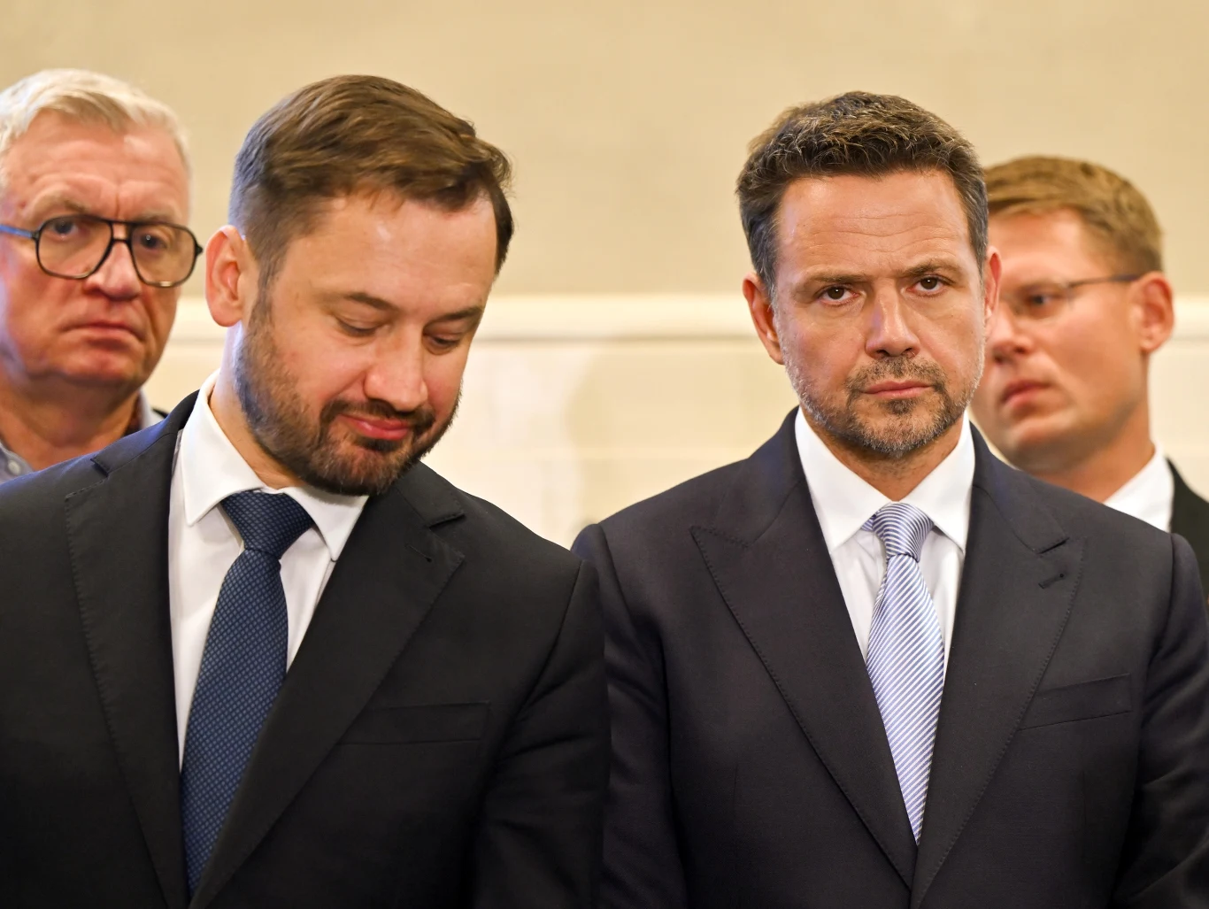 Czterech mężczyzn w garniturach stoi obok siebie, dwóch z nich znajduje się na pierwszym planie, patrząc poważnie przed siebie lub lekko w dół.