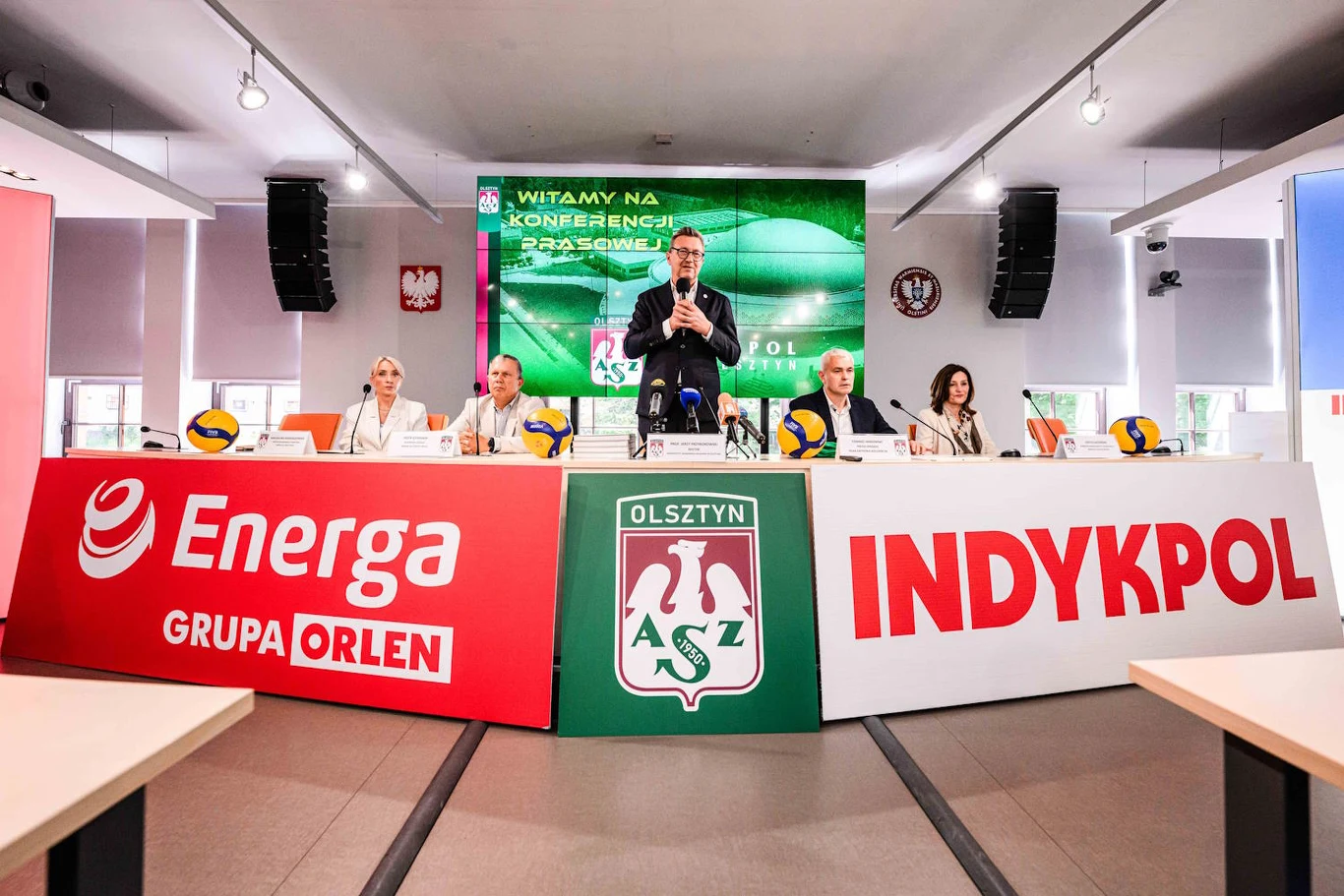 Legendarny polski klub zaczyna nowy rozdział w swojej historii Pięć osób siedzących przy stole konferencyjnym z mikrofonami, tłem z logotypami sponsorów Energa Grupa Orlen i Indykpol oraz herbem klubu AZS Olsztyn, centralnie mężczyzna przemawiający do mikrofonu.