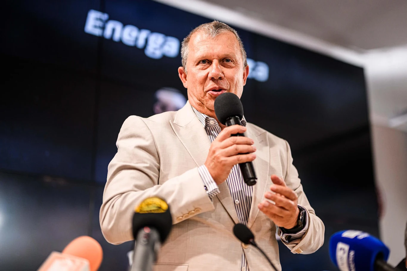 Wiceprezes zarządu Energa SA z Grupy Orlen podczas konferencji prasowej z radością przypomniał, że w zeszłym miesiącu Energa ogłosiła najlepsze wyniki w swojej historii Mężczyzna w jasnej marynarce przemawia do mikrofonu podczas konferencji prasowej, przed nim stoją mikrofony różnych mediów.