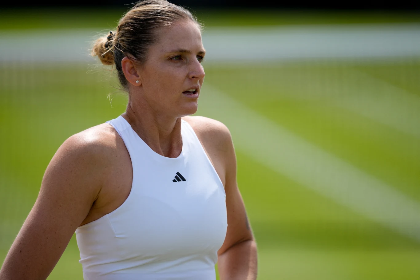 Karolina Pliskova Kobieta w sportowej białej koszulce z logo marki Adidas stoi na tle zielonego kortu tenisowego, z wyrazem skupienia na twarzy.