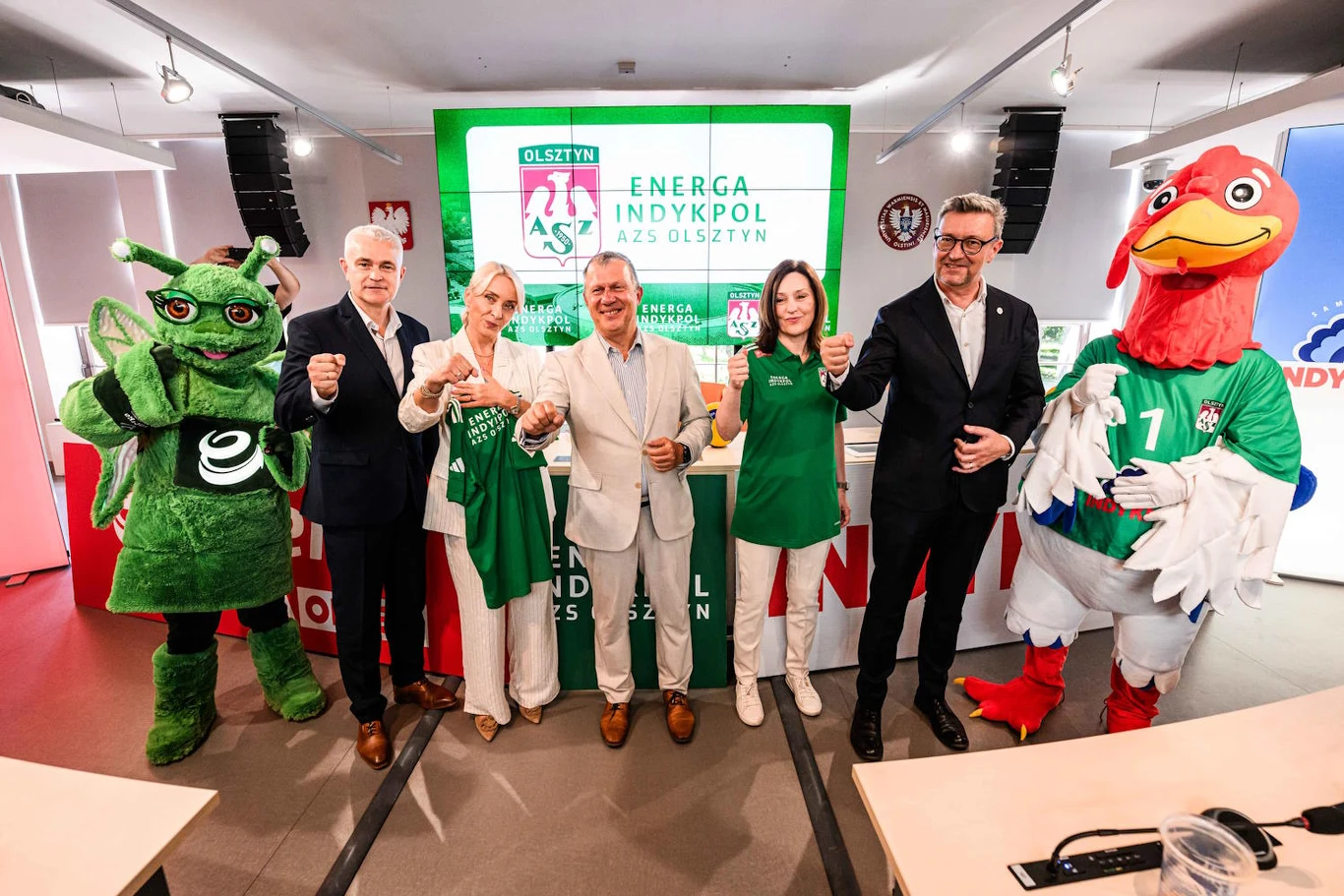 Energa S.A. jako nowy sponsor tytularny liczy na spore sukcesy w nadchodzącym sezonie Grupa sześciu osób, w tym dwie maskotki – zielony owad oraz kogut w stroju sportowym, pozują wspólnie przed ekranem z logo klubu siatkarskiego AZS Olsztyn i sponsora Energa Indykpol, trzymając zieloną koszulkę klubową.
