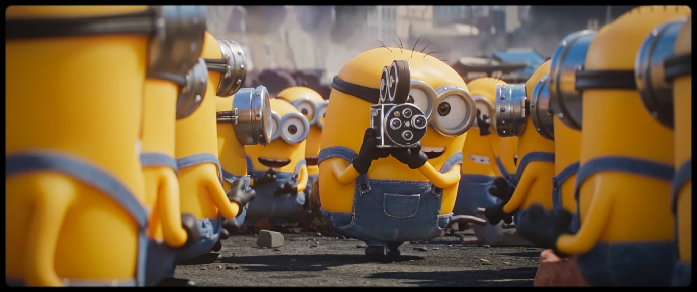 Kadr z filmu "Minionki i straszydła" / UIP Grupa żółtych minionków w niebieskich ogrodniczkach zgromadzona wokół jednego z nich, który trzyma staromodną kamerę filmową i patrzy w obiektyw.