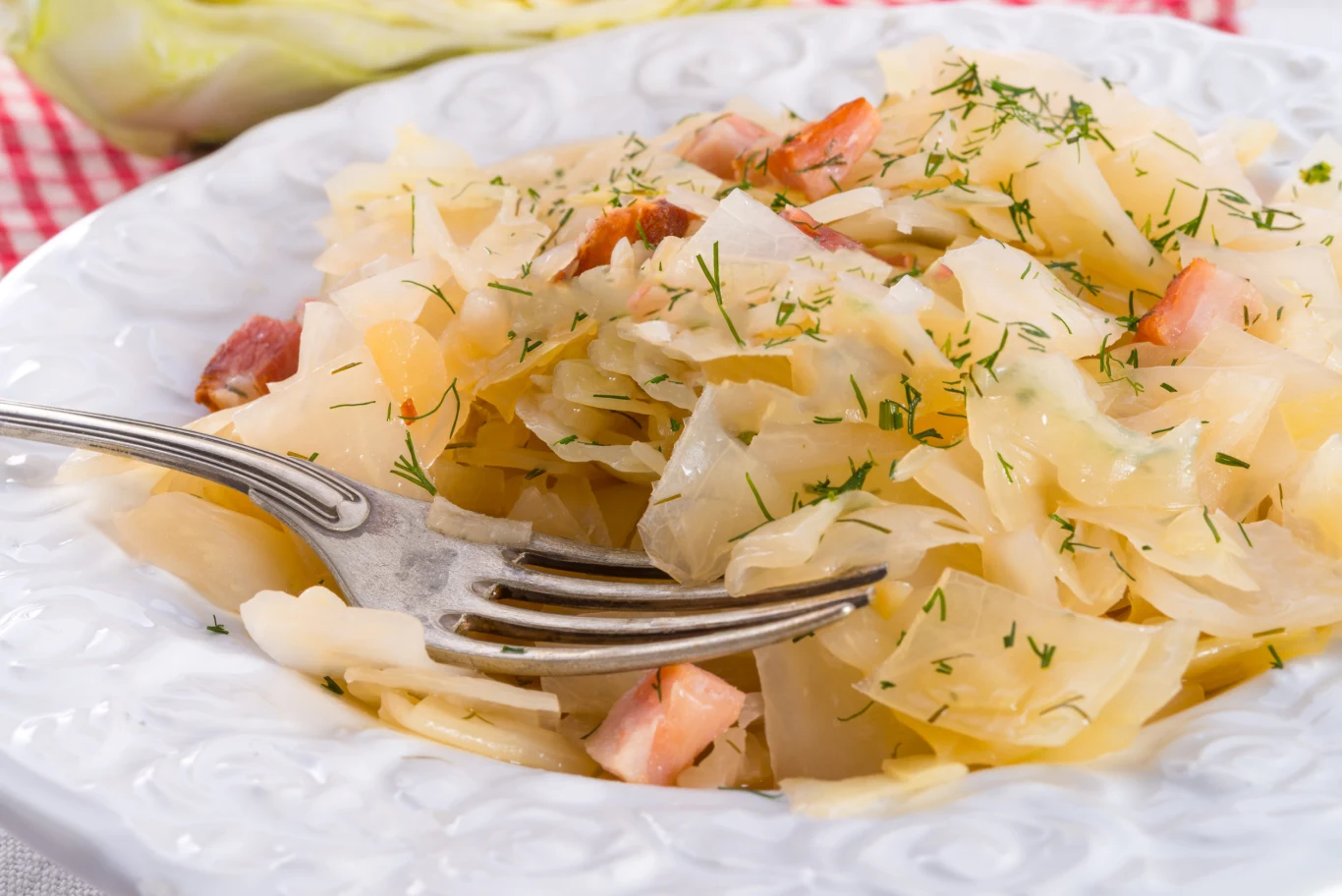 Bigos z młodej kapusty wychodzi delikatny i niezwykle smaczny Bigos z młodej kapusty z boczkiem i koperkiem, podany na białym talerzu z widelcem.