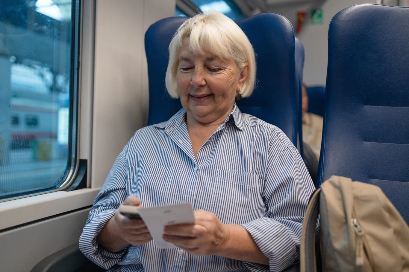 Dzięki rozmaitym zniżkom seniorzy mogą sporo zaoszczędzić na transporcie Starsza kobieta o siwych włosach siedząca na miejscu przy oknie w pociągu, przeglądająca kartkę lub dokument i uśmiechająca się, obok leży beżowy plecak.