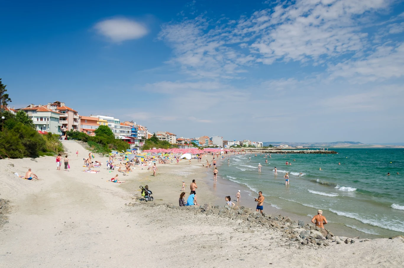 Szeroka, piaszczysta plaża z licznymi wypoczywającymi ludźmi, turkusowe morze oraz budynki i kolorowe parasole w oddali.