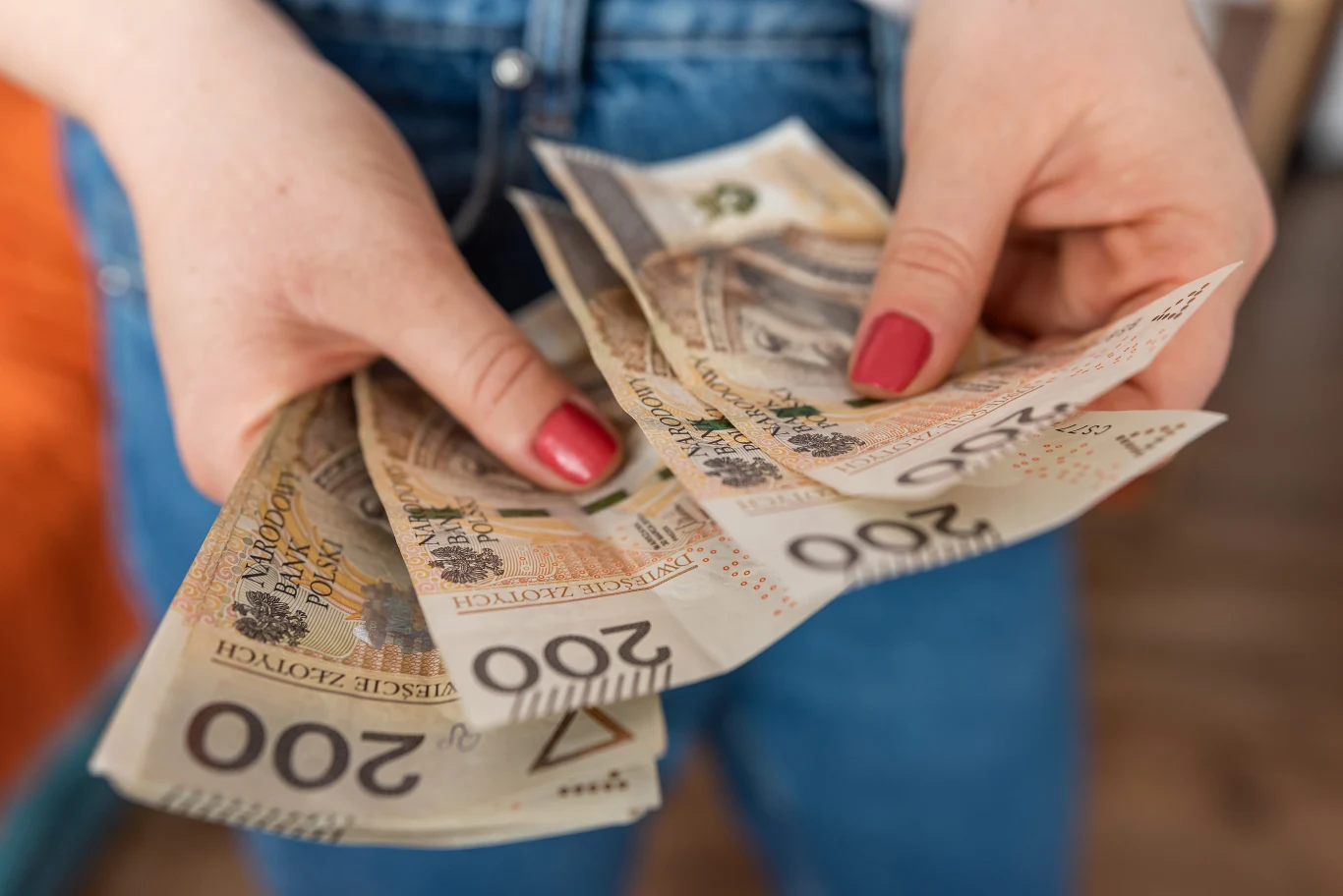 W lipcu możesz liczyć na przypływ gotówki. Wystarczy, że wykorzystasz moc pewnej ciekawej rośliny. Kobieta w jeansach i o czerwonych paznokciach trzyma w dłoniach banknoty o nominale 200 zł.