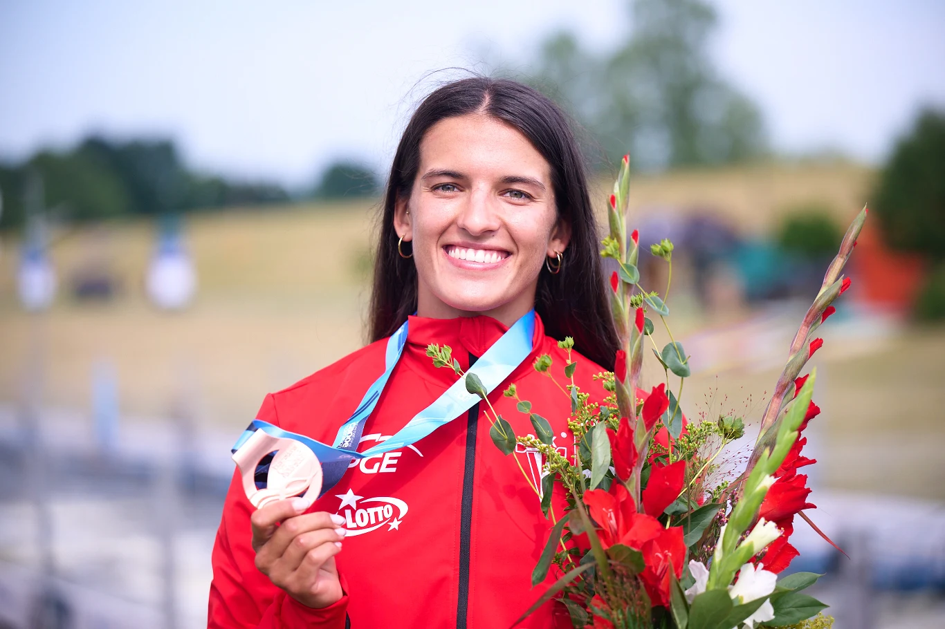 Uśmiechnięta kobieta w czerwonym stroju sportowym trzyma srebrny medal zawieszony na niebieskiej wstążce oraz bukiet czerwono-białych kwiatów.