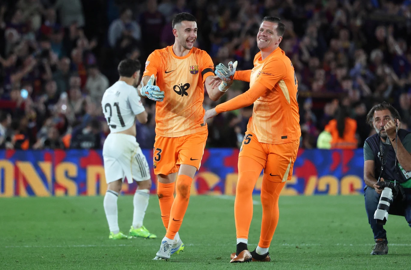 Dwóch bramkarzy w pomarańczowych strojach Barcelony uśmiecha się i trzyma za ręce na murawie stadionu podczas meczu, w tle piłkarz przeciwnika odchodzi, a fotoreporter kuca z aparatem.