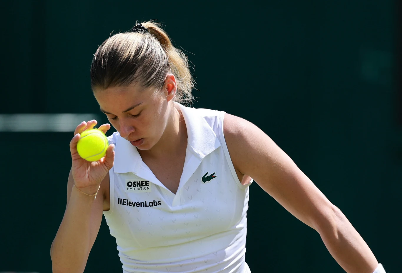 Maja Chwalińska Kobieta ubrana w sportową, białą koszulkę z widocznymi logotypami sponsorów trzyma w ręce żółtą piłkę tenisową, pochylając głowę w skupieniu.
