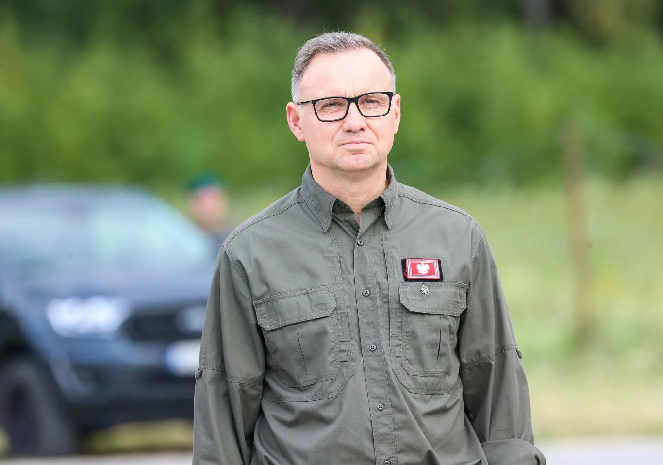Andrzej Duda Andrzej Duda w okularach, w zielonej koszuli z naszywką orła, stoi na zewnątrz, w tle samochód terenowy i drzewa.
