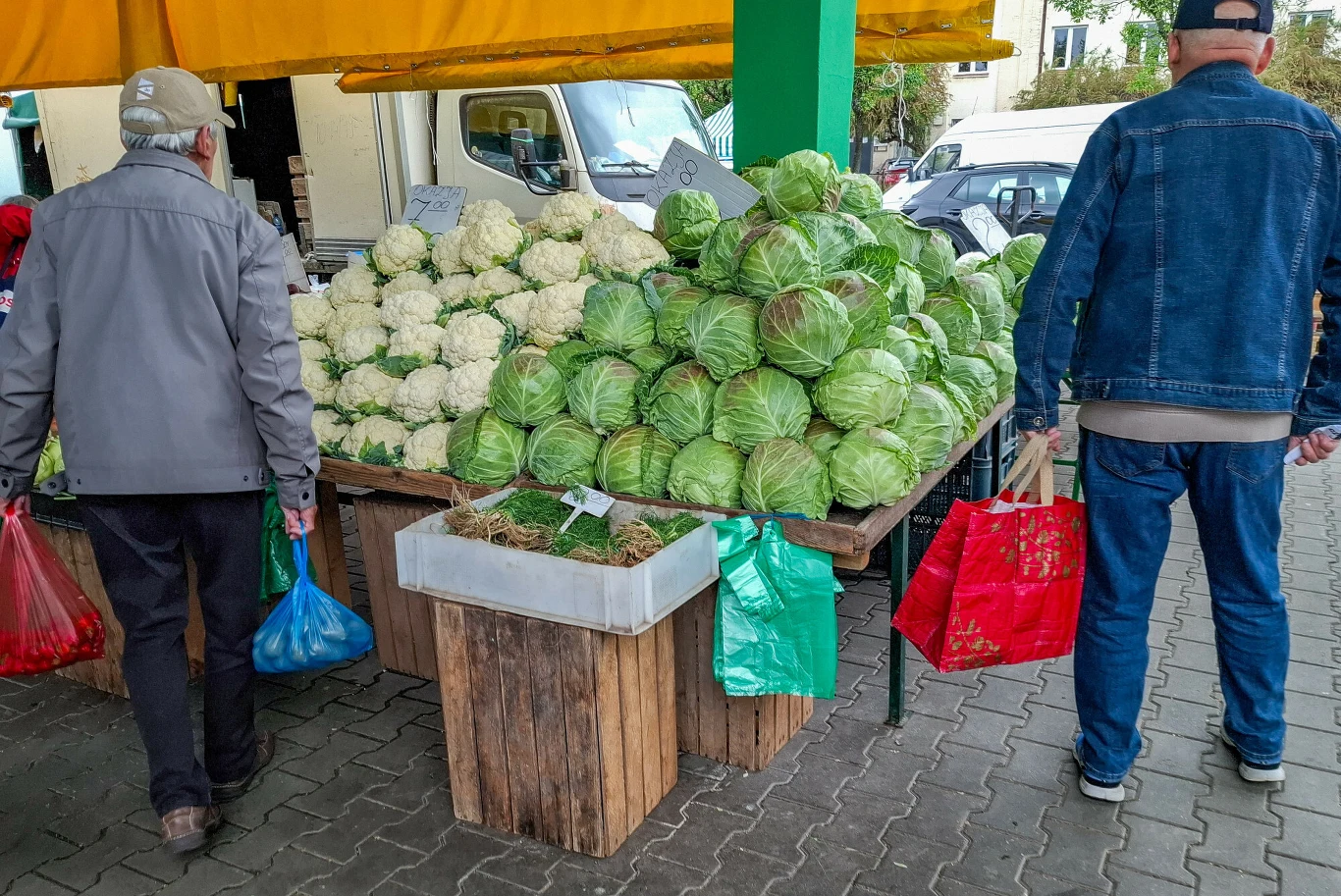 Stoiska z kapustą i kalafiorem na targu, przy których stoją dwaj mężczyźni z zakupowymi reklamówkami.