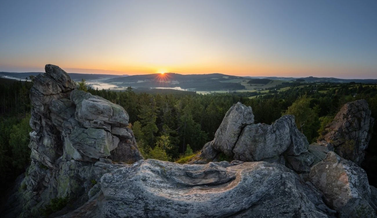 Góry Zdziarskie urzekają malowniczymi widokami na okoliczne lasy i wzgórza Masztywne formacje skalne otaczające naturalne zagłębienie, w tle gęsty las oraz wschodzące słońce nad horyzontem rozświetlającym rozległy krajobraz dolin i wzgórz.