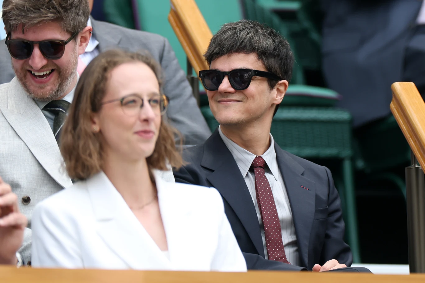 Troje elegancko ubranych osób siedzi na trybunach; dwie z nich mają okulary przeciwsłoneczne i uśmiechają się, atmosfera jest swobodna i radosna.