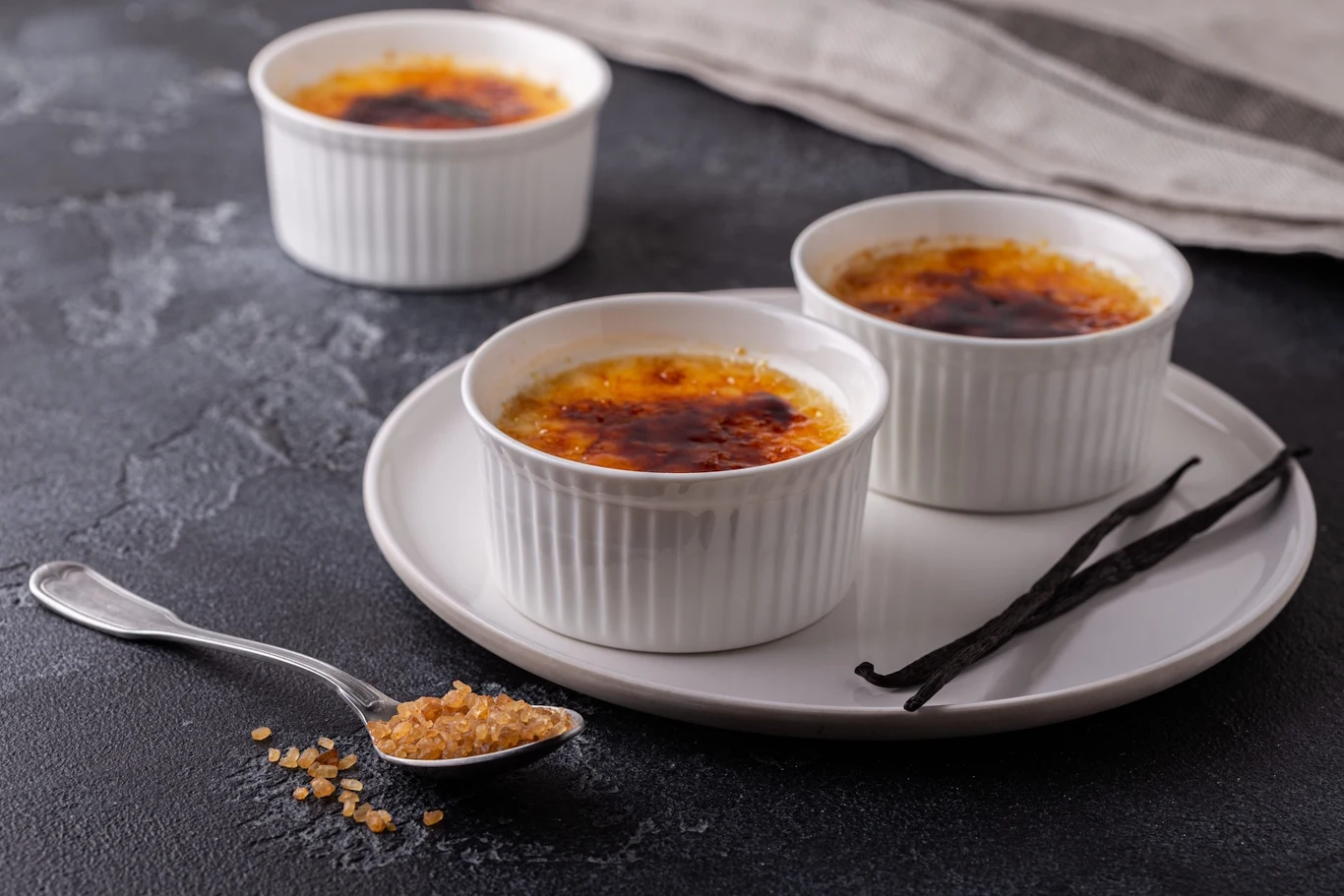 Trzy kokilki z crème brûlée o skarmelizowanej powierzchni ustawione na ceramicznym talerzu, obok laska wanilii i łyżeczka brązowego cukru.