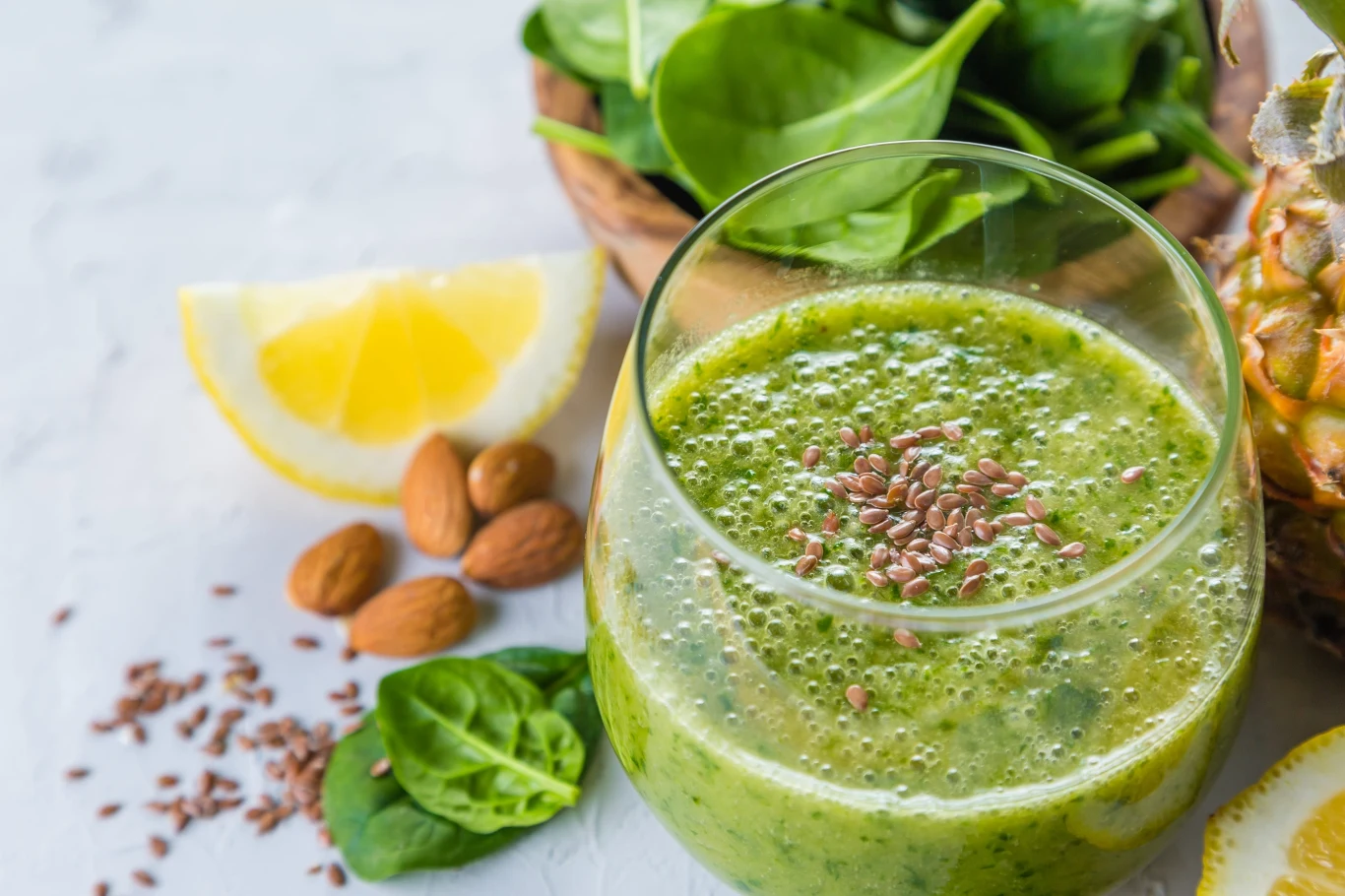 Do zielonego koktajlu możesz dodać także garść ulubionych orzechów, a także sok z limonki bądź cytryny Szklanka zielonego smoothie posypanego siemieniem lnianym, otoczona świeżymi liśćmi szpinaku, kawałkiem cytryny, migdałami i fragmentem ananasa.