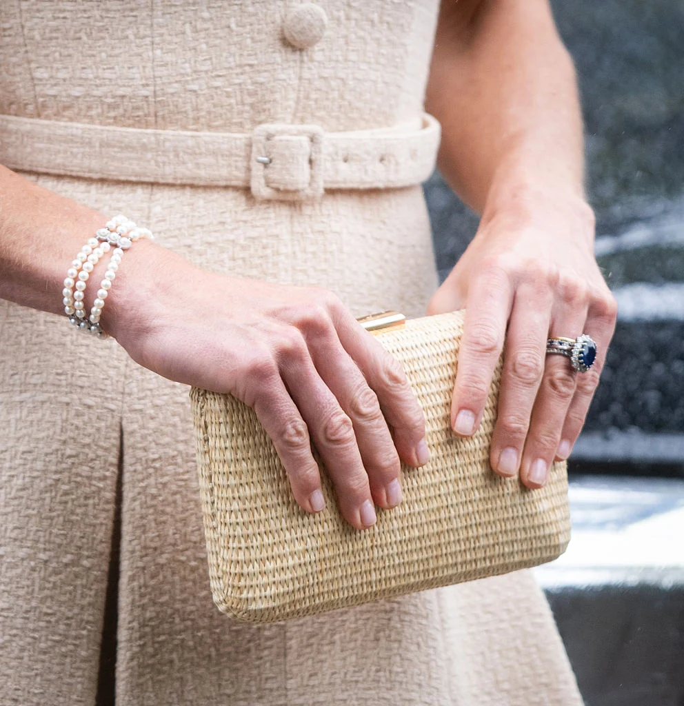 Eleganckie dłonie ozdobione pierścionkiem z dużym niebieskim kamieniem i bransoletką z pereł, trzymające jasną, wiklinową kopertówkę; widoczna jest również beżowa sukienka z materiałowym paskiem i ozdobnymi guzikami.