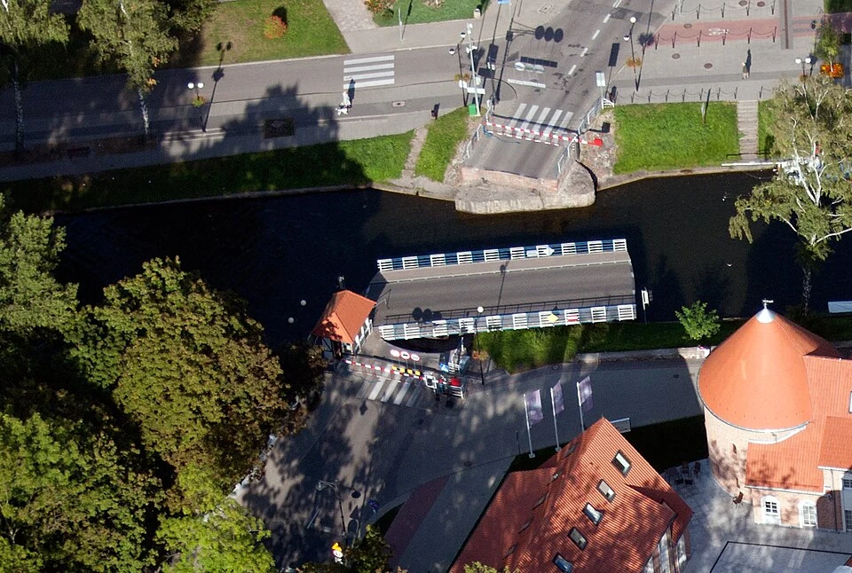 Most zwodzony w trakcie podnoszenia, otoczony zielenią, z widocznym fragmentem rzeki oraz czerwonymi dachami pobliskich budynków.
