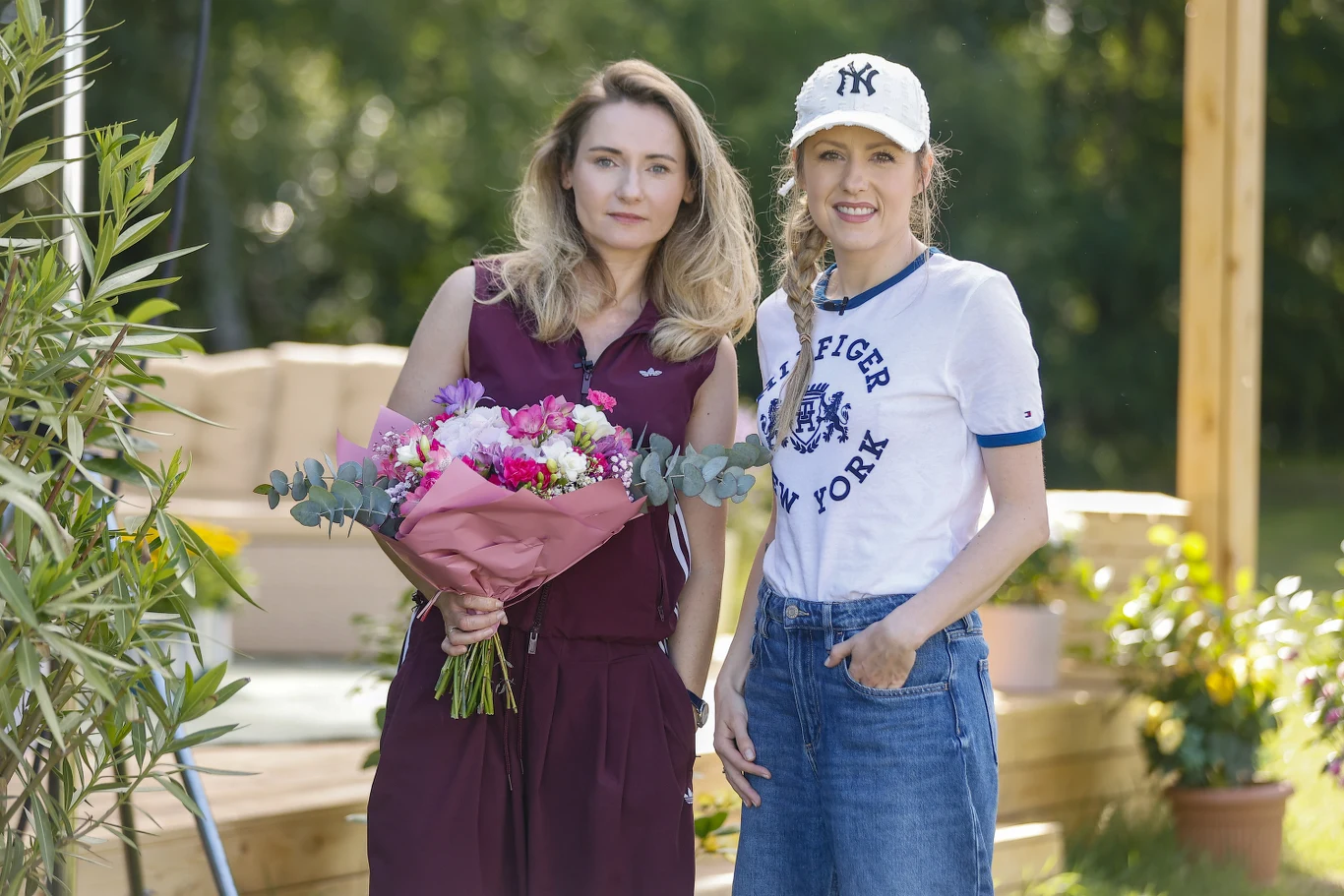 Zuzanna Grabowska i Agata Wątróbska Dwie kobiety stoją obok siebie na drewnianym tarasie otoczonym zielenią; jedna trzyma bukiet kwiatów, druga ubrana jest w białą czapkę z daszkiem i koszulkę sportową.