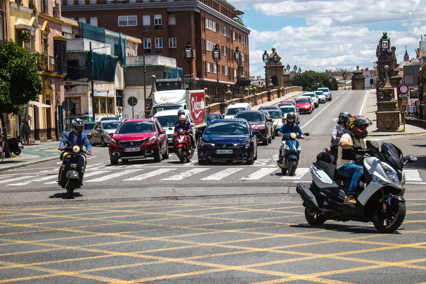 Motocykliści i samochody ustawione przy skrzyżowaniu, gotowi do ruszenia na zielonym świetle, tło stanowią miejskie kamienice i szeroka droga prowadząca na most.