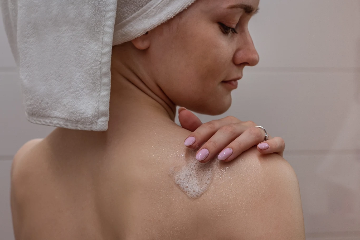 Kobieta z turbanem z ręcznika na głowie delikatnie nakłada żel lub balsam na ramię, skupiając się na pielęgnacji skóry.