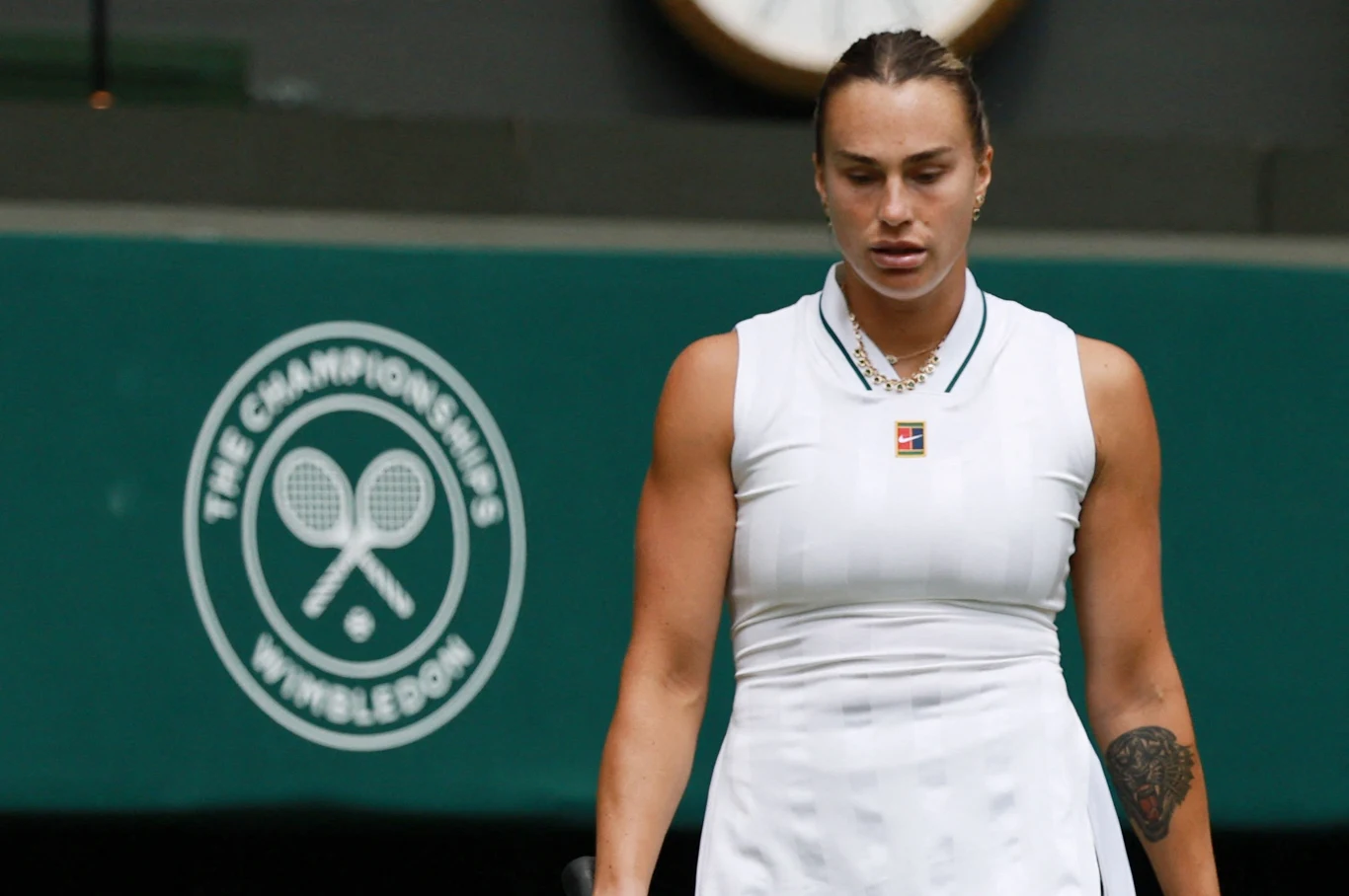 Tenisistka w białym stroju sportowym stojąca na tle zielonej ściany z logo turnieju Wimbledon, z widocznym tatuażem na lewym przedramieniu.
