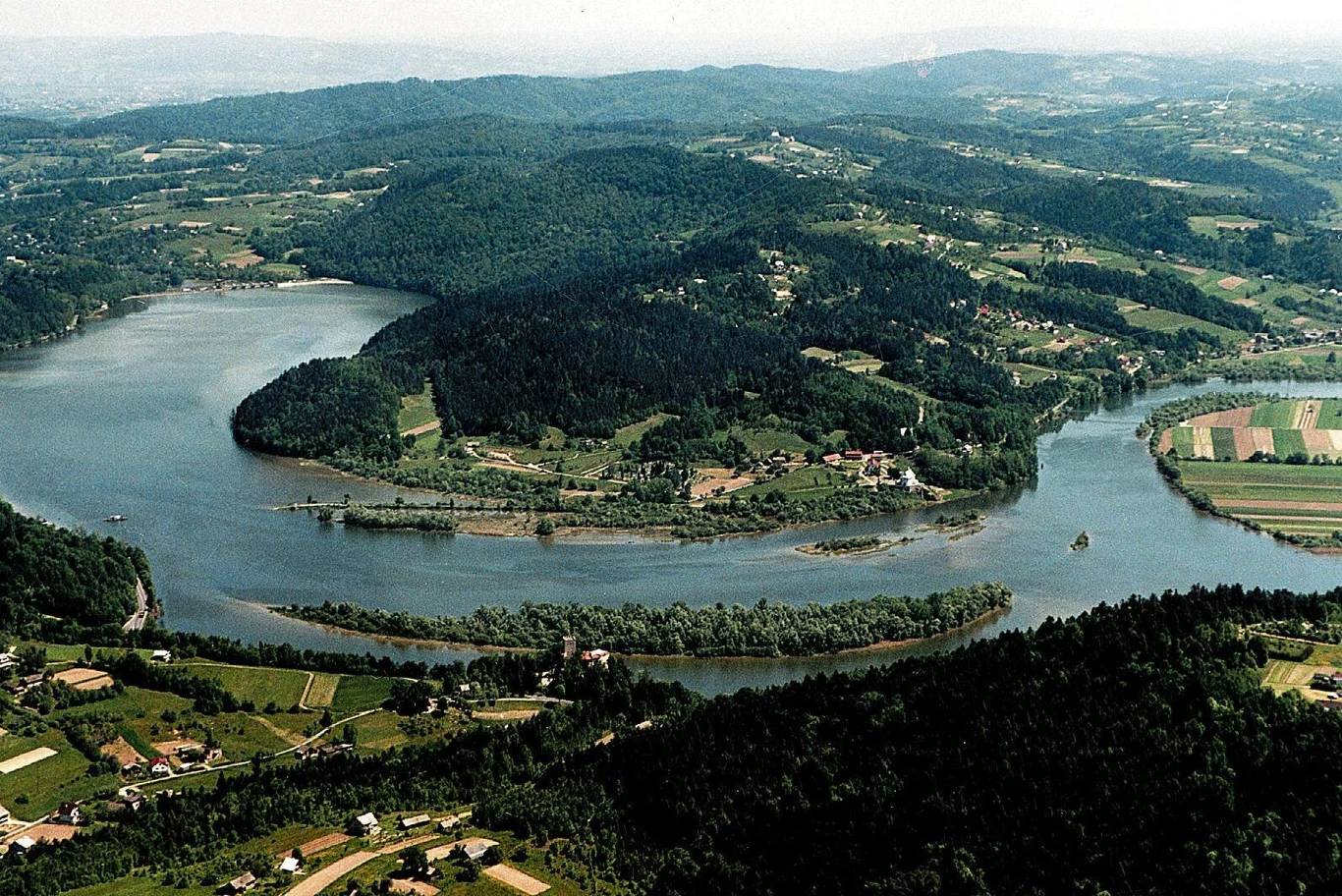 Jezioro Czchowskie, w dole planu zamek Tropsztyn Zakole szerokiej rzeki otoczone zielonymi wzgórzami, polami uprawnymi i rozproszoną zabudową, tworzące malowniczy krajobraz z widocznym pasem drzew na wyspie rzecznej.
