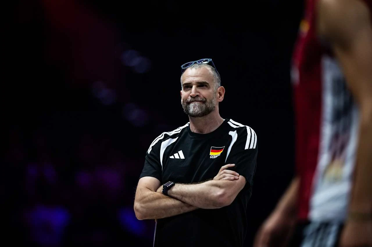 Trener w czarnej koszulce z niemiecką flagą stoi z założonymi rękami i skupionym wyrazem twarzy, obok widoczny fragment sylwetki zawodnika w stroju sportowym.