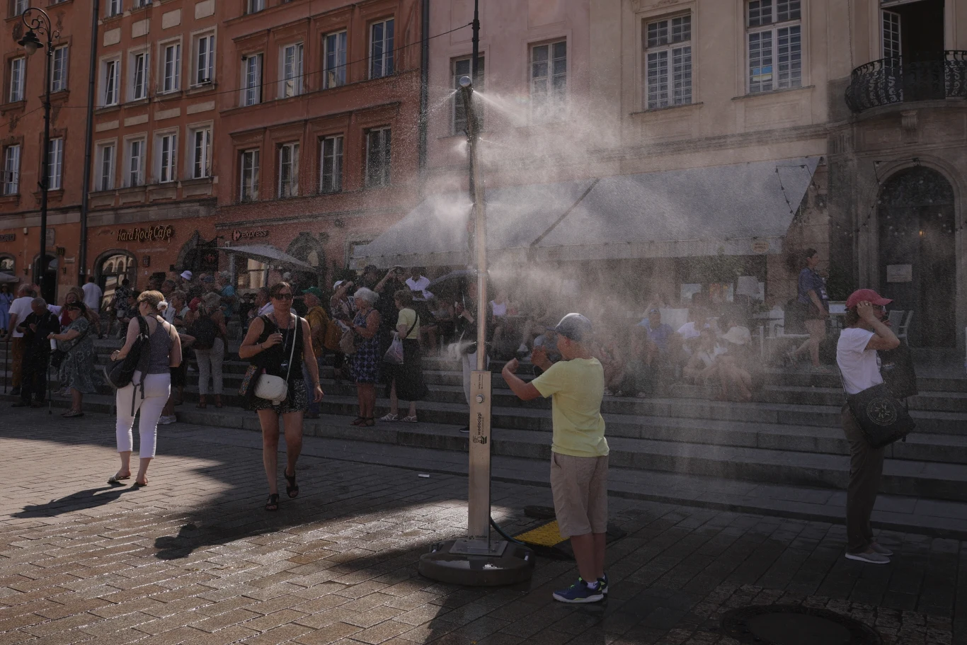 Upały w Polsce osiągają coraz wyższe wartości temperatur Chłopiec w jasnej koszulce pod kurtyną wodną na placu, obok przechodnie i kamienice w tle podczas upałów.